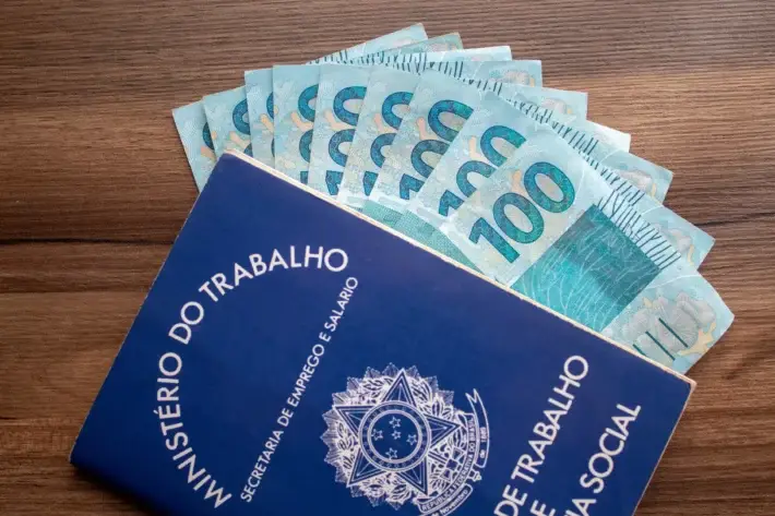 Blue Brazilian government employment booklet (Ministério do Trabalho) with a fan of 100 reais notes emerging from the top.